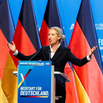 A woman in a dark suit speaks at a podium with both arms raised, in front of German flags and a blue banner reading 'NEUSTART FÜR DEUTSCHLAND. AfD'.