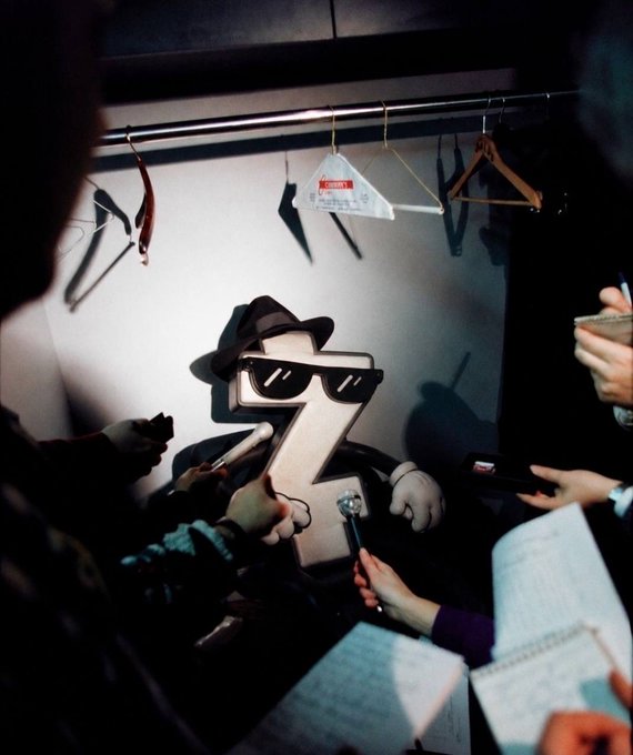 A leather-cased "Z" character wearing a fedora and sunglasses is being interviewed by reporters holding microphones and notepads, playfully visualized as a celebrity altcoin meme. The image cleverly ties to $ZCOIN by depicting the letter "Z" as the cool, newsworthy persona behind the ZEC-inspired token being promoted.