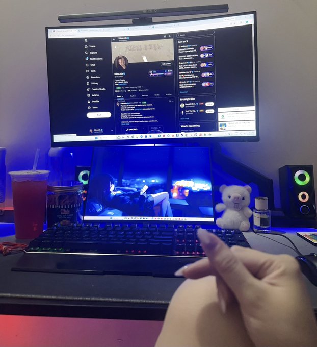 A person's hand resting near a laptop displaying a video game, with a monitor above showing the X (Twitter) social media feed and a small teddy bear on the desk.