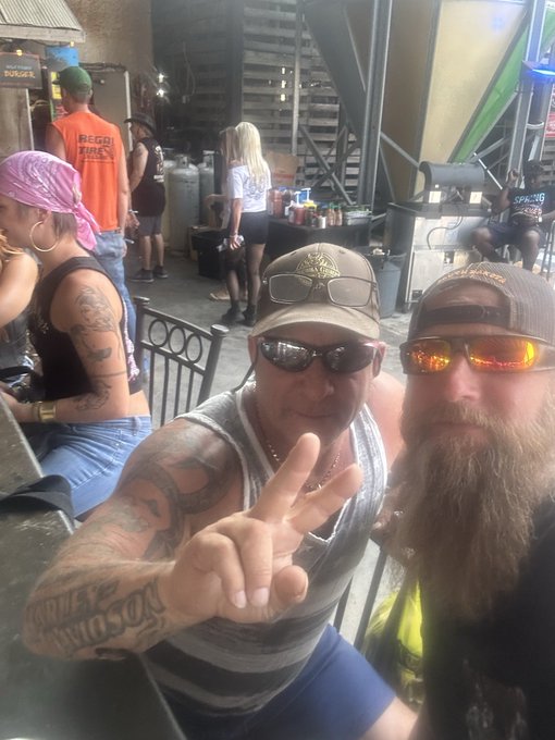 A selfie taken at an outdoor bar or festival showing two men wearing sunglasses and caps; the man on the left makes a peace sign and has a 'Harley Davidson' tattoo on his forearm, with other patrons visible in the background.