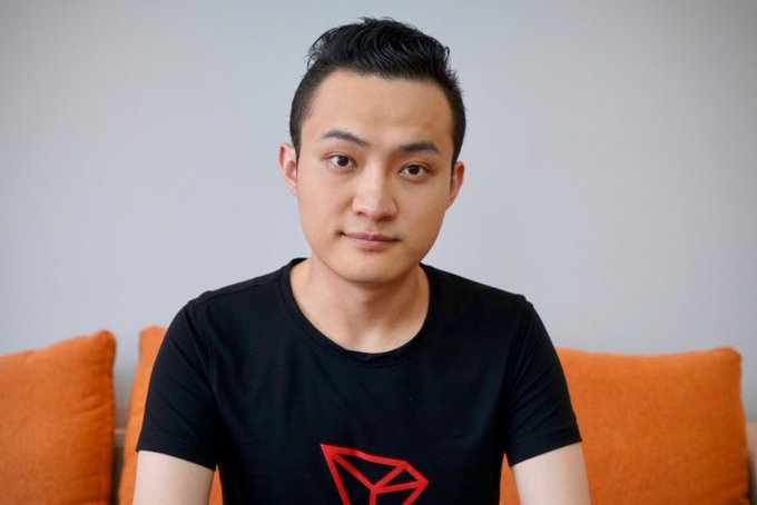 A young man wearing a black t-shirt with a red geometric logo sits on an orange couch against a light gray wall.