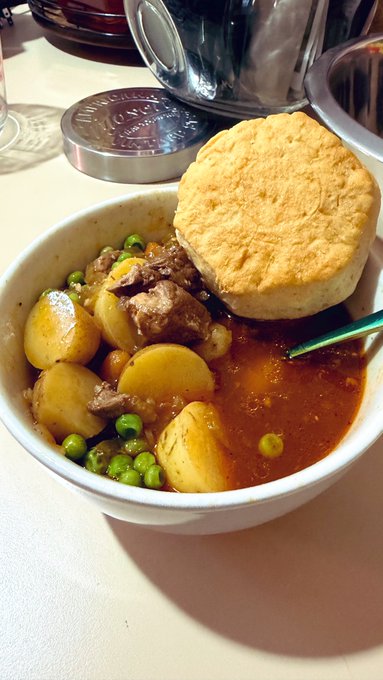 A bowl of beef stew with potatoes, carrots, and green peas, served with a golden-brown biscuit on top, sitting on a white surface.