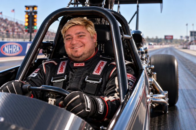 A man in a Simpson racing suit sitting in the cockpit of a drag racing car on an NHRA track, smiling at the camera.