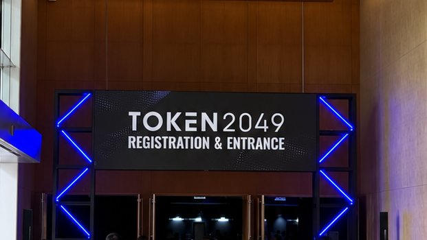 The image shows the registration and entrance signage for TOKEN 2049, a prominent crypto/blockchain conference, illuminated with striking blue neon lighting framing the entryway. The sign directly corresponds to the post's location announcement, marking the attendees' arrival at the event where they're engaging with institutional questions about Ethereum adoption.