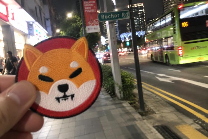 A hand holding a Shiba Inu dog embroidered patch in front of a nighttime street scene in Singapore, with a green '106' bus visible and a 'Rocher Rd' street sign.