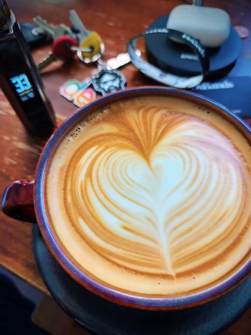 A latte with intricate heart-shaped latte art served in a purple-rimmed cup on a wooden table, with keys, AirPods, and a vape device visible in the background.