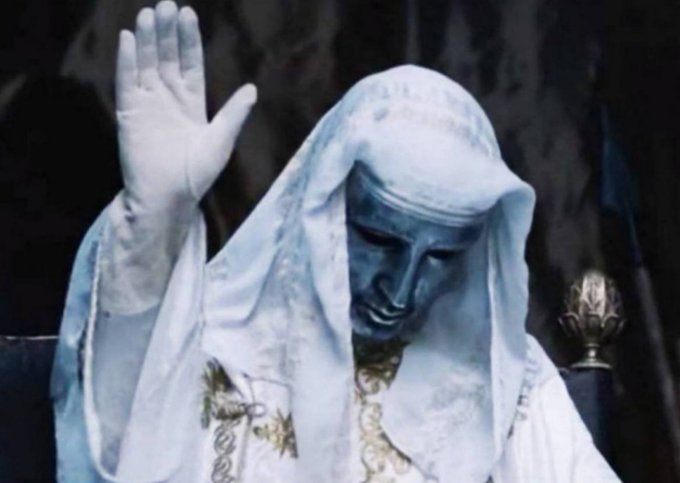 A statue of a figure with blue-tinted skin wearing white robes and a hood, raising one hand with palm facing forward.