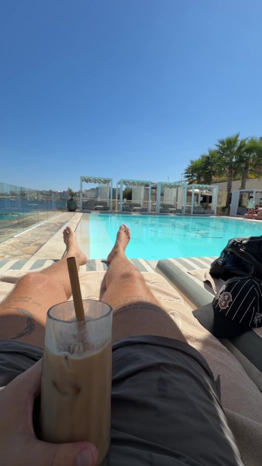 A first-person view of someone relaxing by a swimming pool, holding an iced coffee drink while their bare feet are propped up on a lounge chair.