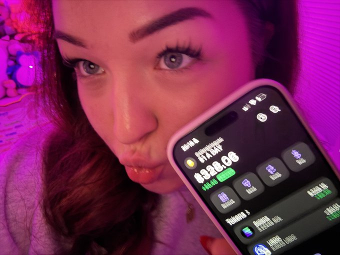 A close-up selfie of a woman under purple/pink lighting, making a pouty expression while holding up a smartphone displaying a money management app showing a balance of '$828.08' for 'Holidaygulaman' with a 'Today +' section visible.