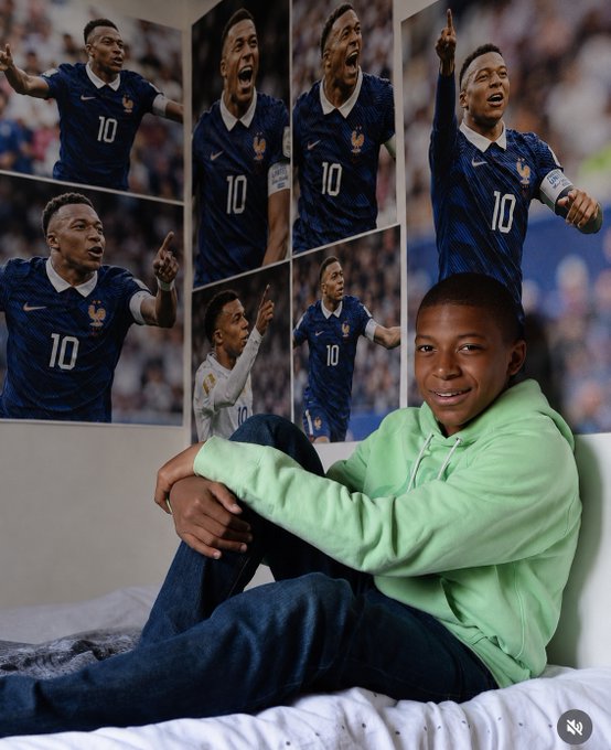 A young boy in a green hoodie sits on a bed, smiling proudly in front of a wall covered with multiple posters of French footballer Kylian Mbappé wearing the number 10 jersey for France's national team. The image ties into the caption by showing that the boy's "poster on the wall" inspiration is his football hero, fitting the idea that every believer—crypto or otherwise—has an idol they look up to.