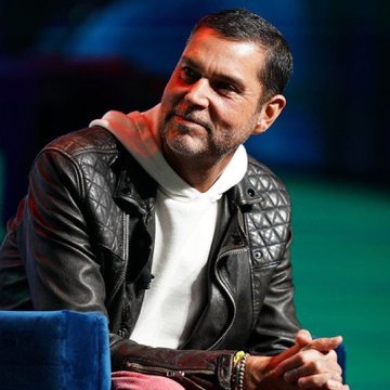 A man wearing a black leather jacket over a white hoodie smiles while seated in a blue chair against a dark green background.