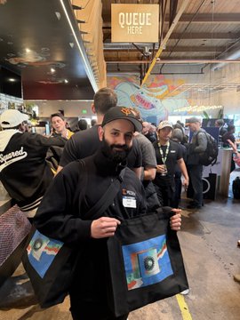 A bearded man holds up a tote bag featuring a colorful record design and "Sound" text, standing in line under a "QUEUE HERE" sign at what appears to be a convention or event. This complements the post celebrating Aesop Solo and his behind-the-scenes contributions to the Miden project.