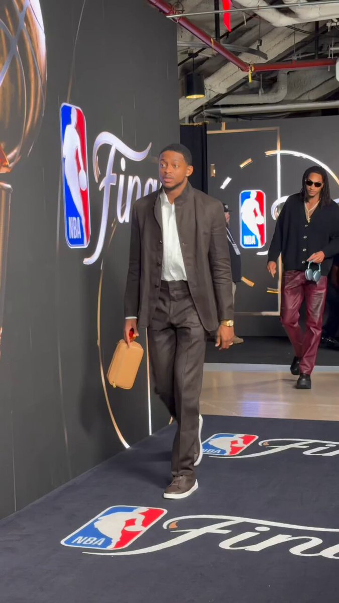 The image shows a man dressed in a suit walking down a hallway, carrying a brown bag. The hallway has a large banner with the words "NHL" written on it, indicating that this scene takes place inside an arena or sports facility related to the National Hockey League (NHL). There are also two other people visible in the image, one of whom is wearing sunglasses and holding a camera.