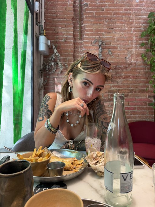 A casual photograph of a tattooed woman with sunglasses on her head sitting at a restaurant table with food, a glass of water, and a large bottle.