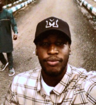 A selfie of a young man wearing a black cap with a white 'M' logo and a plaid shirt, standing on a road.