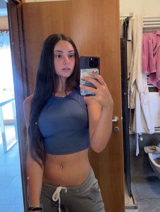 Mirror selfie of a young woman with long dark hair wearing a gray crop top and gray sweatpants, taking the photo in what appears to be a bathroom with a wooden door and hanging towels.