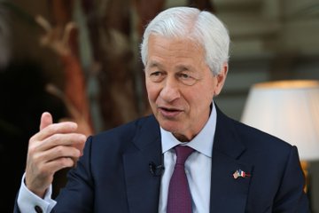 The image shows a man wearing a suit, sitting at a desk with his hand raised as if he's speaking or gesturing. The background features a lamp and a painting, creating an indoor setting for the scene. The text in the image reads "LAST WEEK JAMIE DIMON SAID HE'D FIGHT THE CRYPTO BILL. TODAY COINBASE SAYS every bank wants in." This suggests that there is some sort of discussion or event related to cryptocurrency and its impact on banks, as indicated by the man's raised hand gesture.