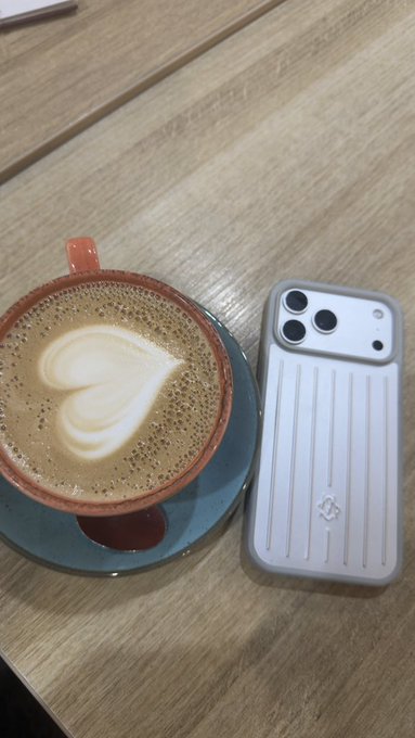 An overhead shot of a wooden table featuring a cup of coffee with heart-shaped latte art in a red and blue ceramic cup, alongside a smartphone in a gray ribbed case.