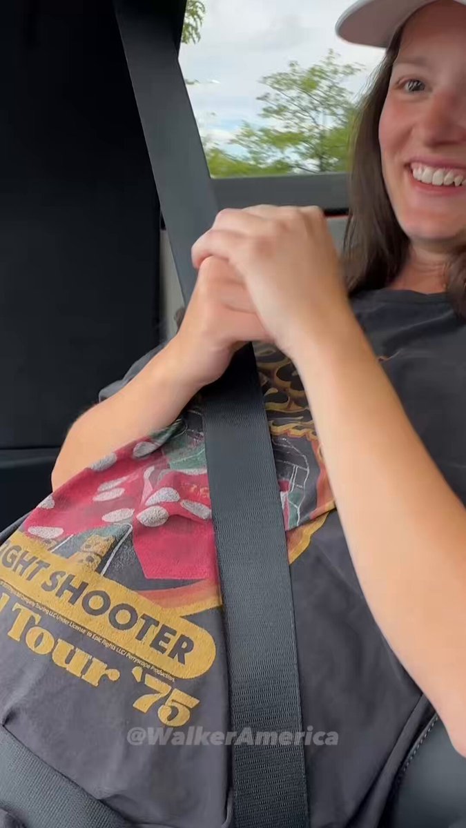The image shows a woman sitting inside a car, smiling at the camera while holding onto her seatbelt with both hands. The background of the photo features a tree outside the window, adding a natural element to the scene. There are also some text elements in the image that read "We are so back #bitcoin is my safe word." and "#bitcoinismysafeword," which seem to be related to Bitcoin or cryptocurrency.