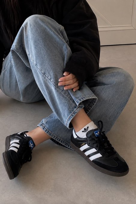 Person sitting on floor wearing baggy blue jeans and black Adidas Samba sneakers with white socks.
