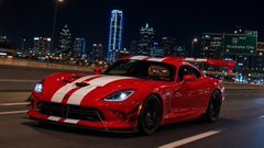 A red sports car with white racing stripes driving on a city street at night with illuminated buildings in the background.