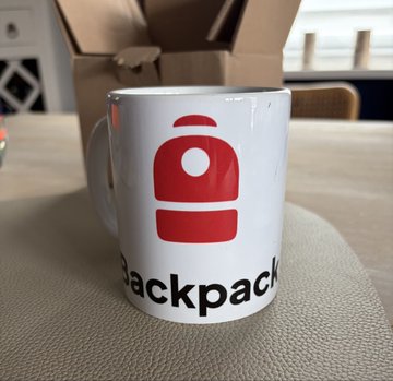A white coffee mug on a table featuring a red robot-like logo and the word 'backpack' in black text, with a cardboard box in the background.