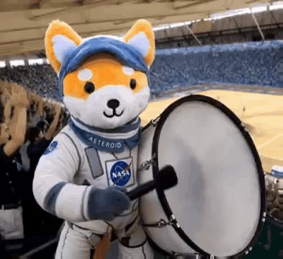 The image features a stuffed animal, specifically a teddy bear wearing a spacesuit, holding a drum set with both hands while standing in front of an audience at a stadium or sports arena. The post also mentions that this event is related to International Day Asteroid plushies and that they are confirmed to be restocking by early September.