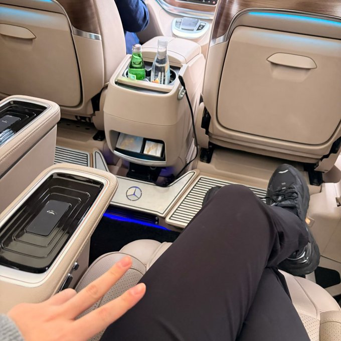 Interior view of a luxury Mercedes-Benz vehicle showing tan leather seats with blue ambient lighting, a center console holding water bottles, and the photographer's legs extended in a relaxed position.
