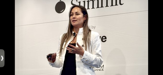 The image features a woman standing in front of a white wall with a sign that reads "SUMMIT" written on it. She appears to be giving a presentation or speech, as she holds up her phone for the camera. The background is black, which contrasts with the white wall and makes the woman stand out more prominently.