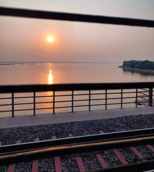 A serene sunset over a calm body of water, viewed through the railing of a bridge or railway with tracks visible below.