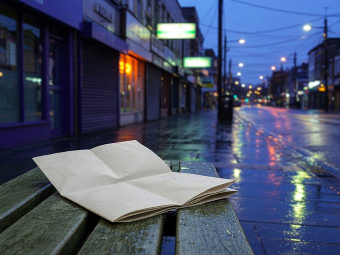 A folded piece of paper rests on a wet wooden bench on a city street at dusk, with neon signs reflecting on the rain-soaked pavement in the background.