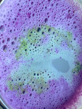 A close-up view of a bubbly, foamy liquid surface with vibrant purple and pink colors mixed with hints of green and yellow.