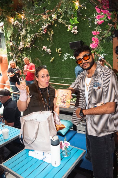 A woman holding a champagne glass and a man wearing sunglasses holding a promotional card, posing together at an outdoor venue with a floral wall in the background.