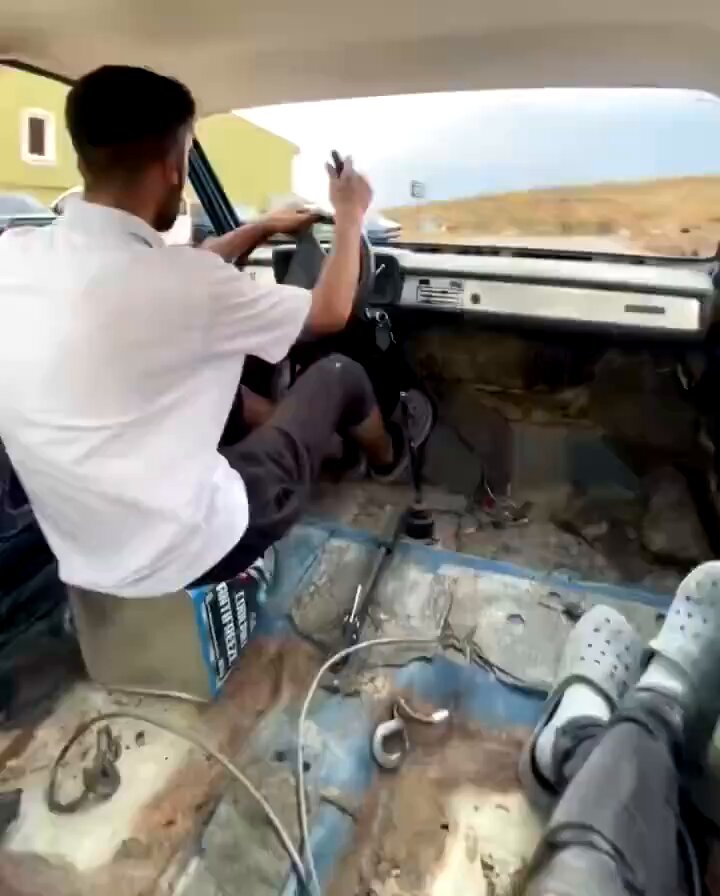 The image shows a man sitting inside an old car with two other people, both wearing flip flops. The scene takes place outside of a house or building. There are also small rocks scattered around the area, indicating that the location may be a rustic or rural setting.