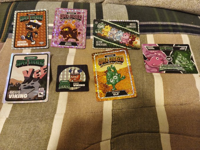 A collection of 'Super Stickers' featuring characters like 'Arrogant Admiral,' 'Bull,' 'Elf,' 'Versatile Viking,' and Pokémon including '6th Dimension' Gengar Hamster and 'Feels/Space/Phantom' boosters, laid out on a plaid blanket.
