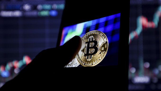 A hand holds a physical Bitcoin token against a backdrop of a digital screen displaying cryptocurrency trading charts and price data. The image visually represents the surge in the crypto market's total value, which surpassed $2.159 trillion in July, gaining over $120 billion that month.