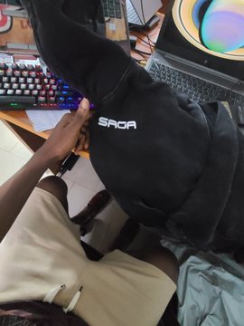 A first-person view shows hands holding a black garment with the text 'SAGA' printed in white, with a keyboard and monitor visible in the background.