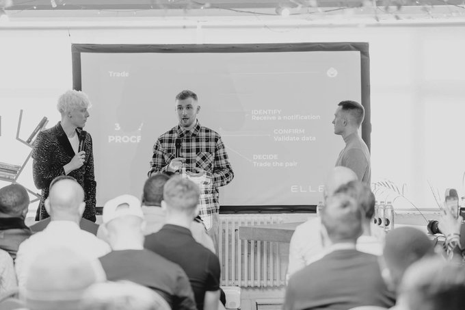 A black and white photograph of three men presenting on stage in front of a projection screen, with the text 'Trade', 'PROOF', 'IDENTIFY', 'CONFIRM', and 'DECIDE' visible behind them.