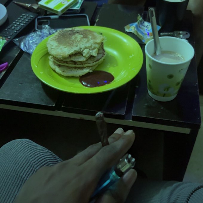 A green plate of stacked pancakes with chocolate sauce and a polka-dot cup of drink on a dark table, with a hand holding a vape in the foreground.