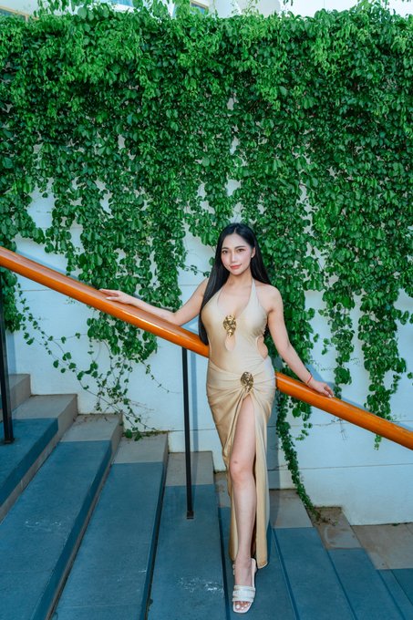 A woman in a beige halter dress with gold accents stands on gray stairs with a wooden handrail, posed against a wall covered in dense green ivy.