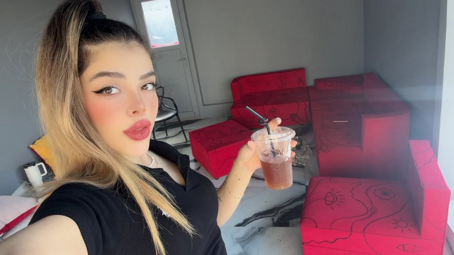 A young woman with a high ponytail takes a selfie while holding a clear plastic cup with a dark beverage, standing in front of a bright red couch with black eye designs.