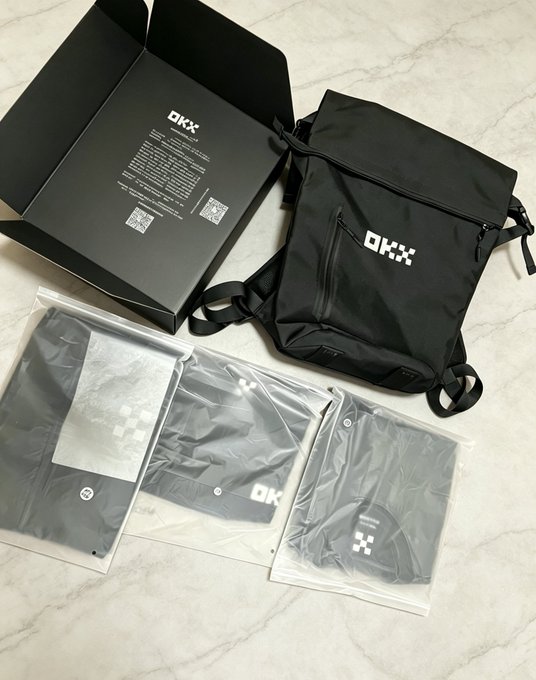 An unboxing photo showing a black OKX-branded box, a black crossbody bag, and several clothing items packaged in clear plastic sleeves.