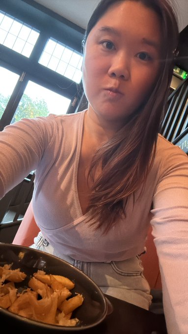 A woman with long dark hair wearing a pink ribbed top takes a selfie at a restaurant, with a bowl of partially eaten pasta visible in the foreground.