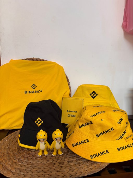 A collection of yellow Binance-branded merchandise, including a t-shirt, bucket hats, and two small yellow figurines, displayed on a woven mat.