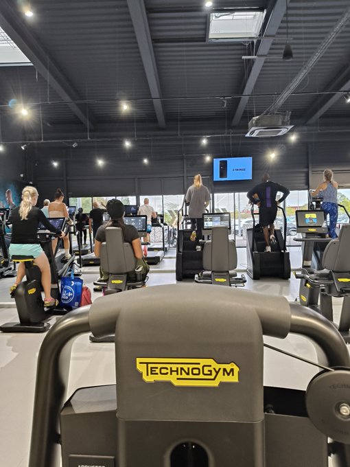 An indoor gym scene shows people using Technogym cardio equipment like stationary bikes and steppers during a group fitness class.