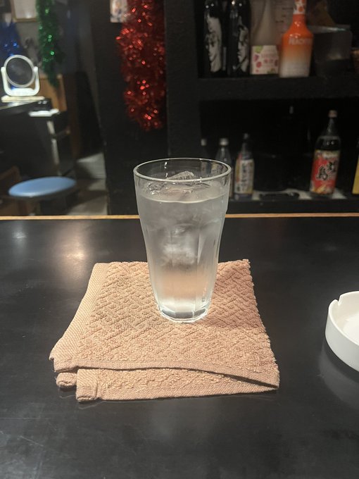 The image features a glass of water sitting on top of a towel, placed on a black countertop. The towel appears to be brown with a patterned design. In the background, there are several bottles lined up against each other, possibly containing various beverages or condiments.