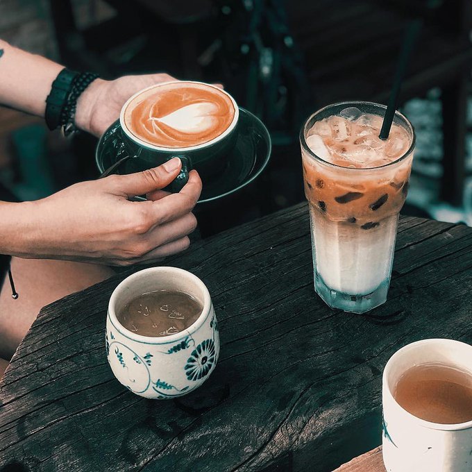 Hands hold a cup of cappuccino with latte art on a wooden table alongside an iced coffee, a small ceramic cup, and another white mug.