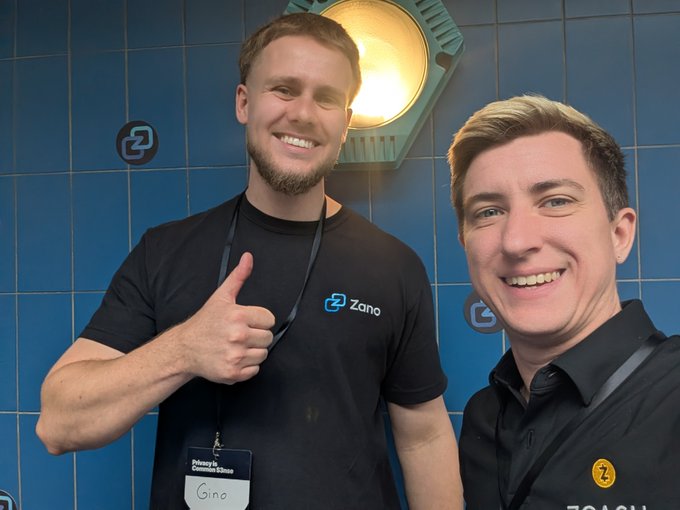 Two smiling men pose for a photo; one gives a thumbs-up and wears a black shirt with a 'Zano' logo, while the other wears a black shirt with a 'Z' logo and a name tag reading 'Gino'.