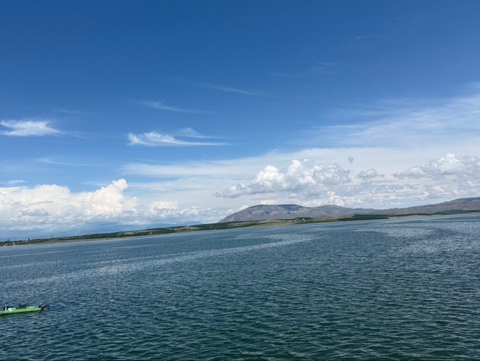 A small boat drifts on a vast, calm sea beneath a brilliant blue sky scattered with fluffy clouds, with distant mountains forming a serene horizon. The open, expansive water and tranquil atmosphere evoke the freedom and possibility often associated with the "GM" (grand mission) journey ahead.