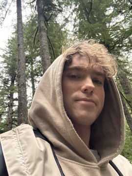 A selfie of a young man wearing a tan hoodie with the hood up, standing in a forest setting with tall trees behind him.
